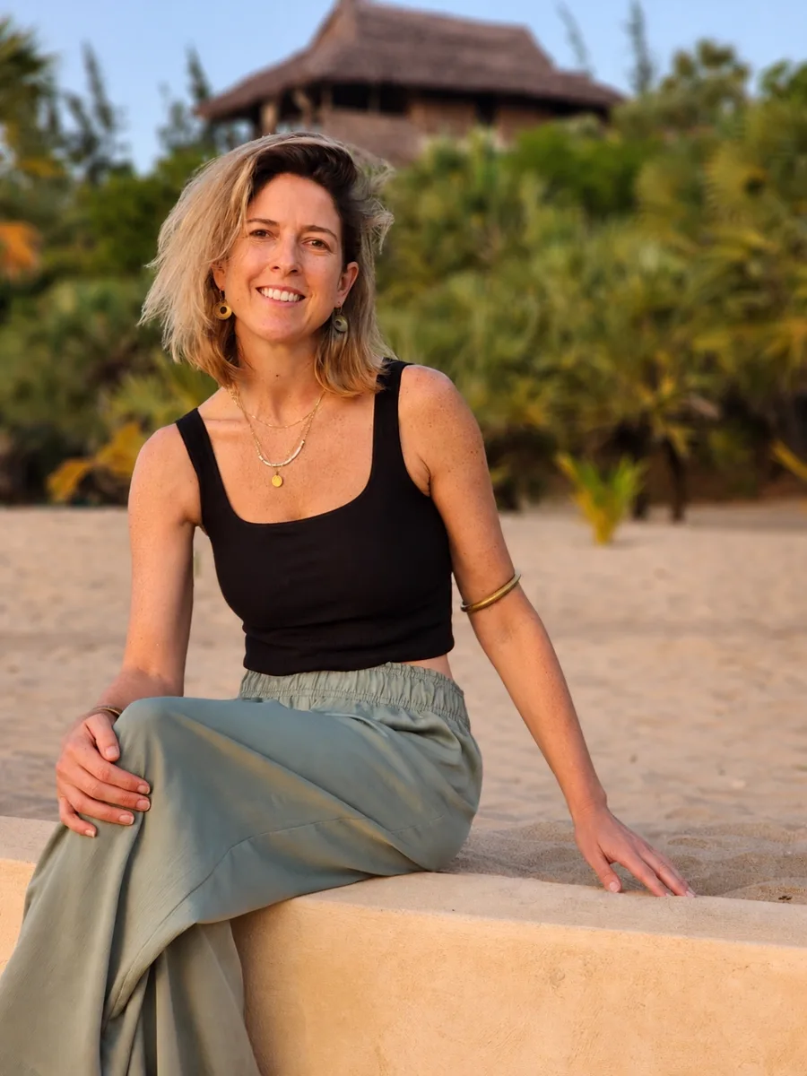 Anna Campbell seated on a sunlit beach wall in front of a thatched lodge and tropical greenery